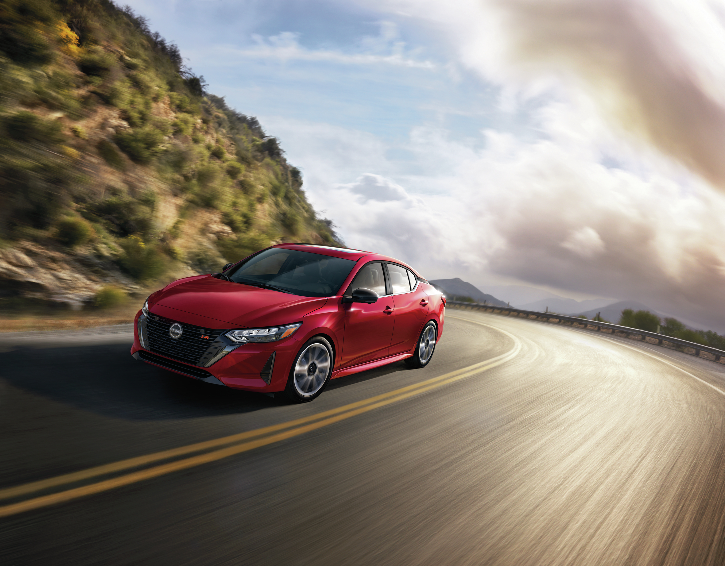 2025 Nissan Sentra SR in Scarlet Ember cruising along a scenic mountain road with a dramatic sky in the background.